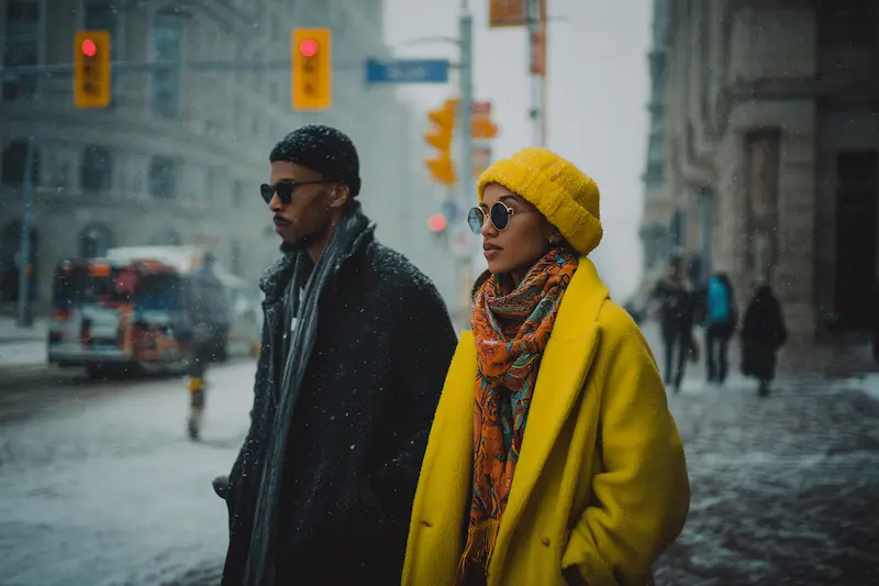 A stylish couple walking side by side in winter, the woman in a bold yellow coat and the man in dark tones, visually representing the contrasting personalities in Aries and Capricorn relationships