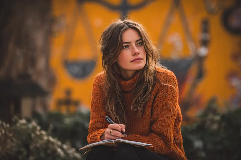 A thoughtful young woman in an orange sweater writing in a notebook outdoors, with a symbolic Libra scales mural in the background, representing balance and astrology