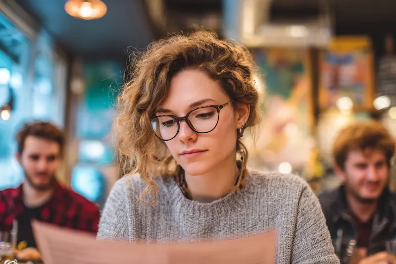 A young woman wearing glasses looking thoughtful and indecisive while reading a restaurant menu, symbolizing Libra’s tendency to struggle with decision-making.